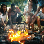 Group of mixed age group of people relaxing by a bonfire and preparing marshmallow candy